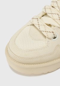 Beige sneaker met een gestructureerde mesh bovenkant en suede accenten, met geweven veters in een kruispatroon en een gevoerde zool.