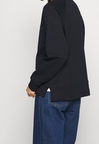 Felpa nera con vestibilità rilassata, polsini a costine e spacchi laterali. Abbinata a jeans a gamba larga blu per un look casual.
