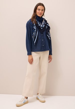 Camicia in denim navy con chiusura a bottoni, abbinata a una sciarpa blu a fantasia. Pantaloni crema a gamba larga e sneakers bianche con accenti blu chiaro.