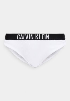 Białe bikini z czarnym elastycznym paskiem w talii z napisem "CALVIN KLEIN" białymi wielkimi literami na jednolitym jasnym tle.