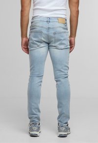 Light blue slim-fit jeans with a faded texture, five pockets, and a tan leather patch on the back waistband, paired with gray sneakers.