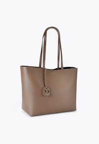 Borsa a mano in pelle marrone con due manici lunghi, design minimale e un'emblema decorativo in oro appeso a una tracolla. Texture liscia.