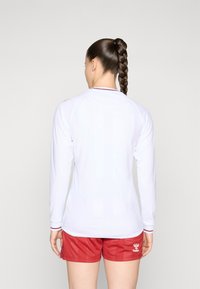 Langærmet hvid sports top med røde accenter og rund halsudskæring, parret med røde shorts. Glat tekstur, tætsiddende design.