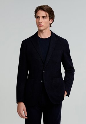 Blazer oscuro texturizado con solapas de muesca, dos bolsillos frontales y cierre de un botón, usado sobre una camiseta de cuello redondo oscura.