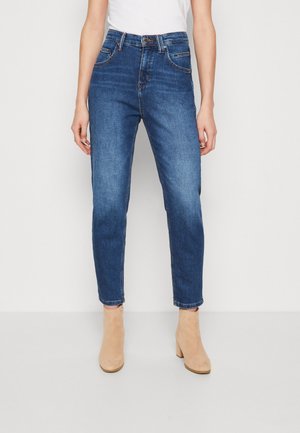 Frau trägt hochtaillierte mittelblaue Jeans und beige Stiefeletten, steht vor einem einfarbigen weißen Hintergrund.