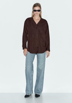 Camicia marrone con bottoni, collo a V, maniche lunghe e tasche frontali, abbinata a jeans a gamba larga di colore blu chiaro e scarpe nere.