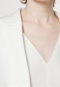 Bílý blazer s klopou, zobrazený s světlým topem s výstřihem do V pod ním. Hladká textura látky a detaily přizpůsobeného střihu jsou viditelné.