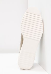 Semelle d'une chaussure beige avec des rainures horizontales appuyée contre un mur blanc, montrant le motif de la bande de roulement et le design du talon plat.