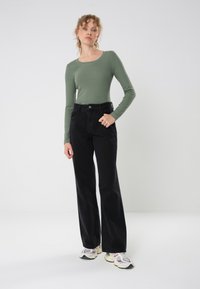 Maglietta verde a coste a maniche lunghe con scollatura a barco abbinata a jeans neri a vita alta e a gamba larga, esposti su uno sfondo neutro.
