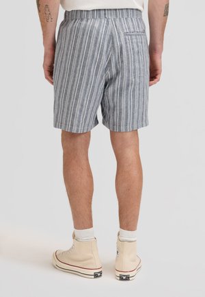 Mann, der graue und weiße vertikal gestreifte Shorts und beige High-Top-Sneaker trägt, von hinten vor einem einfarbigen Hintergrund gezeigt.