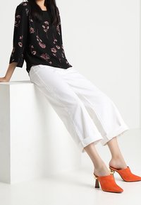 Schwarze Bluse mit floralem Muster und Dreiviertelärmeln, weiße Cropped-Hosen und orangefarbene Mules mit goldenen Akzenten. Glatte Textur für das gesamte Outfit.
