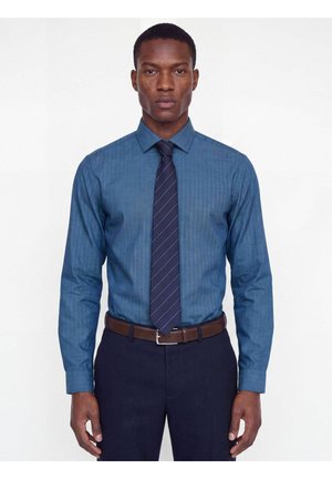 Homme portant une chemise à rayures bleues, une cravate à rayures foncées, une ceinture marron et un pantalon foncé, debout devant un fond blanc uni.