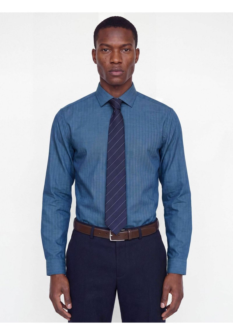 Homme portant une chemise à rayures bleues, une cravate à rayures foncées, une ceinture marron et un pantalon foncé, debout devant un fond blanc uni.