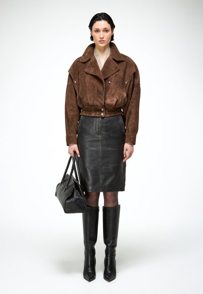 Femme portant une veste courte en daim marron, une jupe en cuir noire jusqu'aux genoux, des bottes noires montantes jusqu'aux genoux, tenant un sac à main en cuir noir.