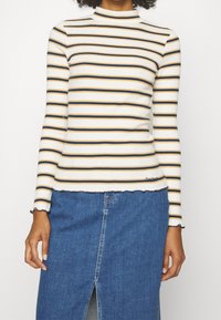 Langärmliges gestreiftes Oberteil in Weiß mit Marineblau- und Senfgelbstreifen, mit hohem Kragen und gewelltem Saumdetail. Kombiniert mit einem Jeansrock.