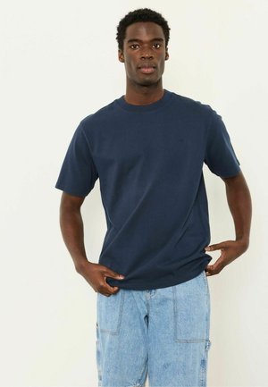 Junger Mann trägt ein navyblaues Kurzarmhemd und hellblaue Jeansshorts, steht vor einem schlichten weißen Hintergrund.
