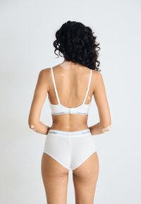 Kvinne med krøllete hår og vitiligo-flekker som modellerer hvit Calvin Klein-bh og høytlivsunderdel, stående mot en ensfarget lys bakgrunn.