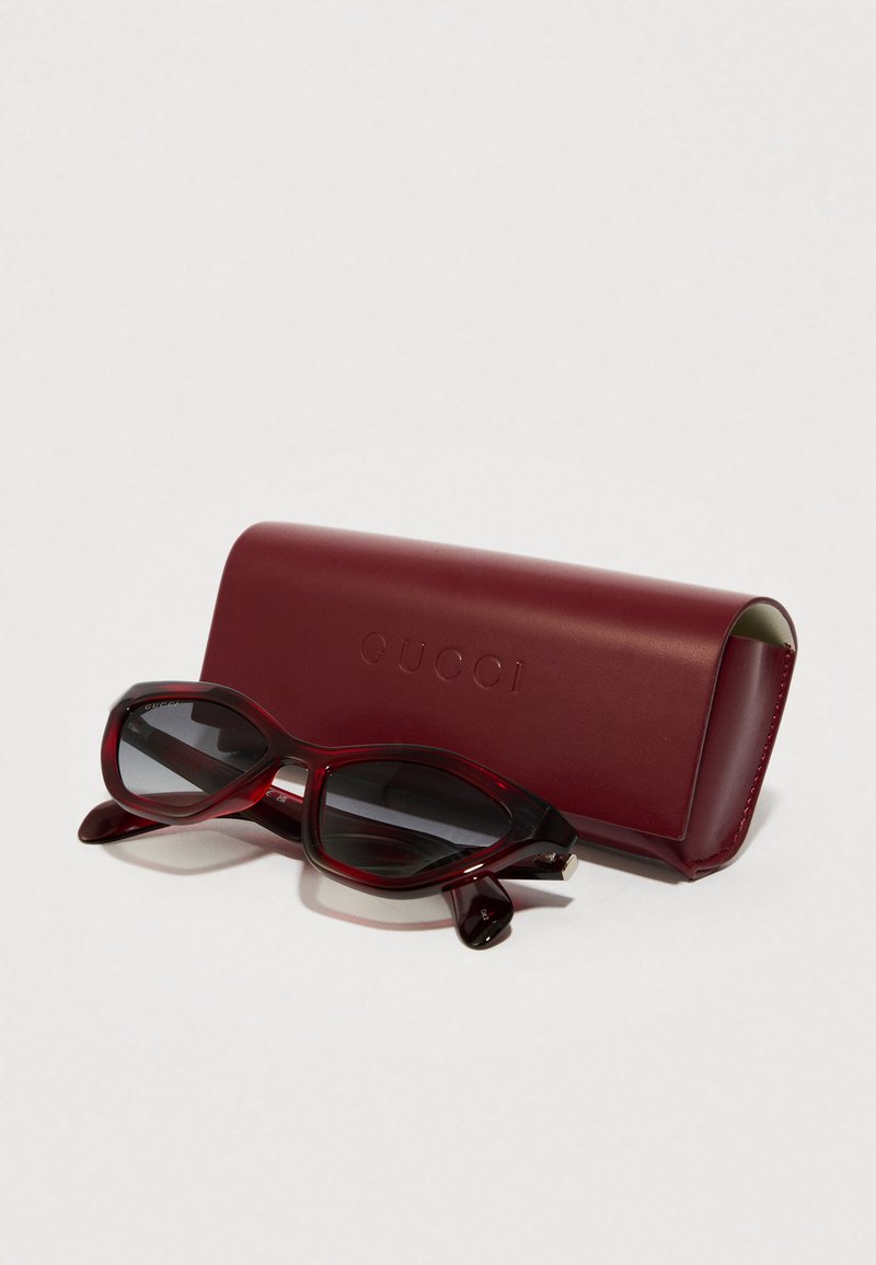 Gucci burgundy zonnebril met een slank ontwerp, donkere lenzen en logo-details, geplaatst naast een bijpassend leren etui.