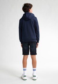 Hanorac bleumarin cu o glugă mare, pantaloni scurți din denim închis, adidași albi cu accente verzi și șosete albe cu dungi albastre, vista din spate.