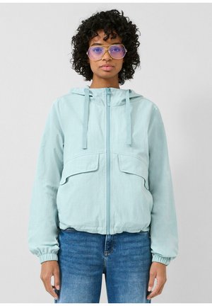 Frau mit lockigem Haar, die eine hellblaue Kapuzenjacke mit Reißverschluss, übergroße blau getönte Brille und blaue Jeans trägt, steht vor einem einfarbigen Hintergrund.