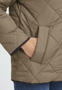 Quilted olive green jacket with a black trim at the cuffs and a side pocket, featuring a smooth texture and diamond pattern.