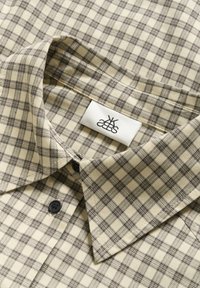 Camicia a quadri beige con sottili linee blu scuro, dotata di colletto a revers e bottoni neri. È visibile la texture del tessuto intrecciato. Etichetta cucita all'interno.