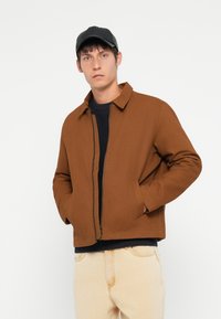 Brown zippered jacket with a classic collar and side pockets, paired with a navy sweater and light-colored pants. Black cap worn.