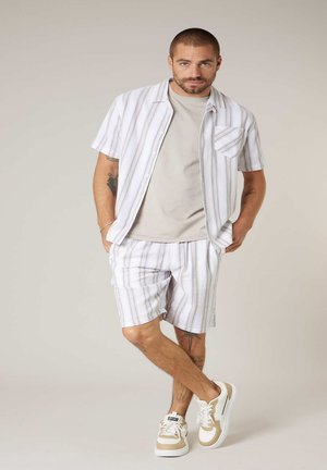 Mann, der ein beigefarbenes T-Shirt, ein weißes kurzärmliges Hemd, passende gestreifte Shorts und weiße Sneaker trägt, mit gekreuzten Beinen steht.