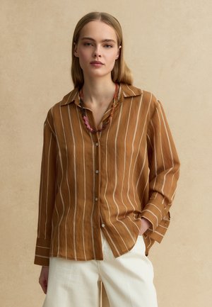 Mujer con camisa marrón de botones y rayas verticales blancas, pantalones blancos y un collar trenzado multicolor, posando frente a un fondo beige.