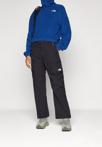 Niebieski polar z wysokim kołnierzem, zestawiony z czarnymi spodniami cargo z dużymi kieszeniami. Szare buty trekkingowe dopełniają stylizację.