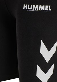 Zwarte stof met wit "HUMMEL"-logo en "SPORTSWEAR SINCE 1923" tekst boven twee witte naar beneden gerichte chevron pijlen.