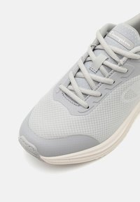 Chaussure de sport grise avec un upper en mesh, des panneaux texturés, une pointe arrondie et une semelle blanche contrastante avec un logo sur la languette.
