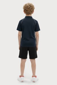 Bērns ar īsiem cirtainiem matiem stāv vērsts projām, ģērbies tumši zilā polo krekla, melnu šortus un baltās kedas ar sarkanām detaļām.