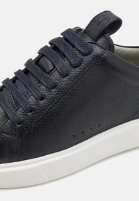 Marineblå lærersneaker med teksturert overdel, flate lisser og hvit gummisåle. Har perforeringer for ventilasjon og en liten hæltab.