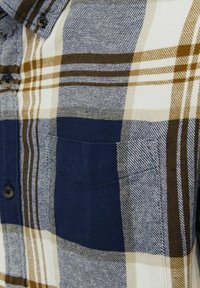 Camisa de botones con un patrón de cuadros en azul, crema y marrón. El material es tejido y cuenta con un solo bolsillo en el pecho, del lado izquierdo.
