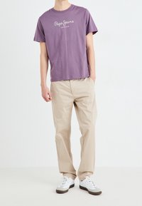 T-shirt viola a maniche corte con logo bianco, abbinato a pantaloni beige larghi e scarpe da ginnastica bianche con accenti grigi.