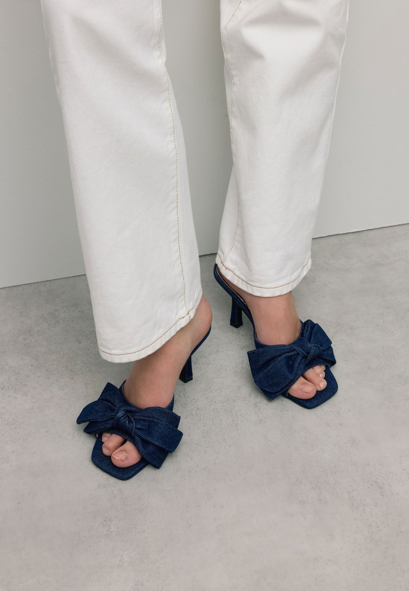 Denim muiltjes met een vierkante neus, 10 cm hoge stilettohak en grote strikdetail op de wreef, in een donkerblauwe kleur. Witte broek zichtbaar.