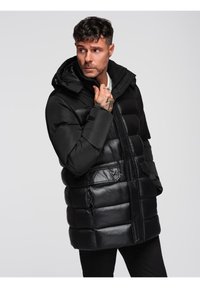 Hombre con un abrigo de invierno negro acolchado y capucha, ajustando el cuello, mirando de lado contra un fondo claro y liso.
