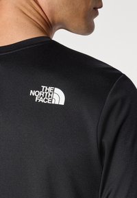 Cămașă sportivă neagră dintr-un material texturat, cu un logo alb "The North Face" pe spate, aproape de guler.