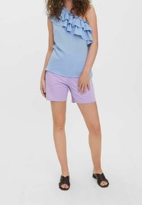 Light blue one-shoulder top with ruffled detail, paired with lavender shorts. Model wears black slip-on sandals with patterned design.