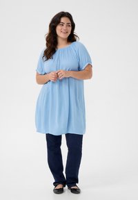 Blouse bleu clair à manches courtes avec encolure froncée, associée à un jean bleu foncé et des ballerines noires. Texture lisse, coupe décontractée.