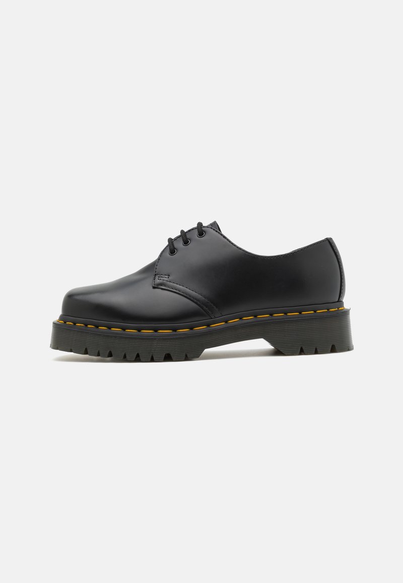 Zapato de cuero negro con cordones, suela gruesa con ranuras y costura amarilla a lo largo del borde de la suela, mostrado en perfil lateral sobre fondo blanco.