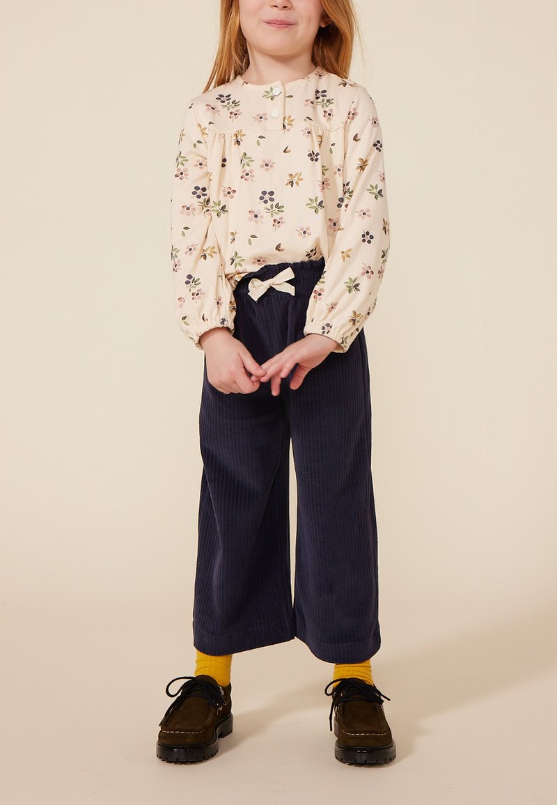 Crèmekleurige blouse met lange mouwen en bloemenprint, gecombineerd met donkerblauwe corduroybroek, voorzien van een strikdetail in de taille. Zwarte schoenen, gele sokken.