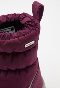Botte isolée violette avec un haut froncé, tissu lisse, une sangle texturée et un accent de logo métallique. Une doublure douce est visible à l'intérieur.
