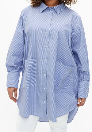 Loose-fitting, long-sleeve blue and white vertical striped shirt dress with front buttons and two large front pockets worn with white pants.