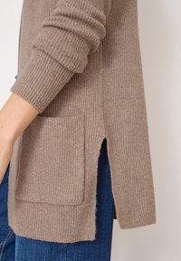 Cárdigan marrón de canalé con una abertura lateral y un bolsillo en la parte delantera, combinado con denim azul, que muestra una construcción de punto texturizado.