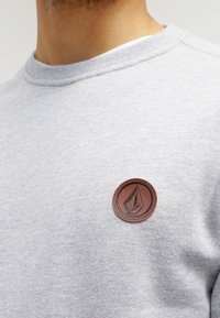 Nahaufnahme einer Person, die einen hellgrauen Sweatshirt trägt, auf dessen Brust ein kleines rundes braunes Lederpatch mit einem geometrischen Logo zu sehen ist.