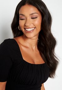 Sort ribbet top med en firkantet udskæring og korte ærmer, med samlet tekstur og tætsiddende design. Håret er langt og bølget.