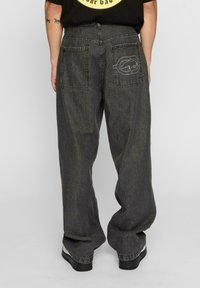 Svart baggy denimbukser med løs passform, to store påsydd lommer på baksiden, og diskrete logo-sømdetaljer.