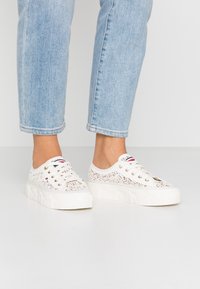 Persona che indossa sneakers platform bianche con motivo di pizzo e occhielli dorati, abbinate a jeans cropped azzurri, su uno sfondo semplice.